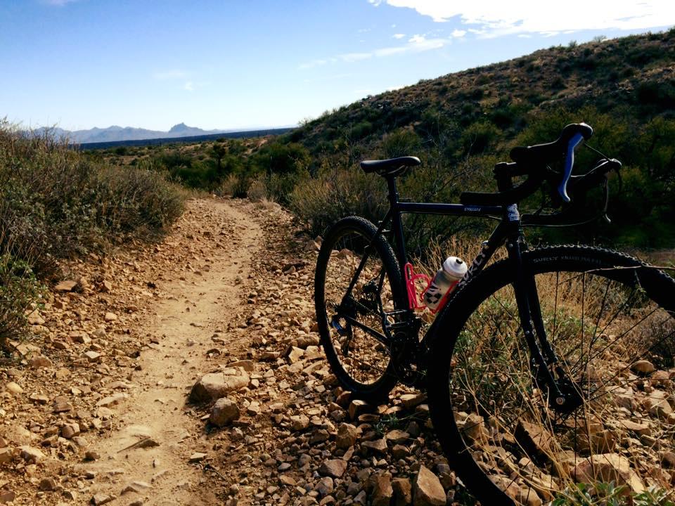Surly Surly Straggler: A black bicycle is parked on a rocky, dirt trail surrounded by sparse vegetation, with mountains visible in the distance under a blue sky. The bike features a water bottle attached to the frame.