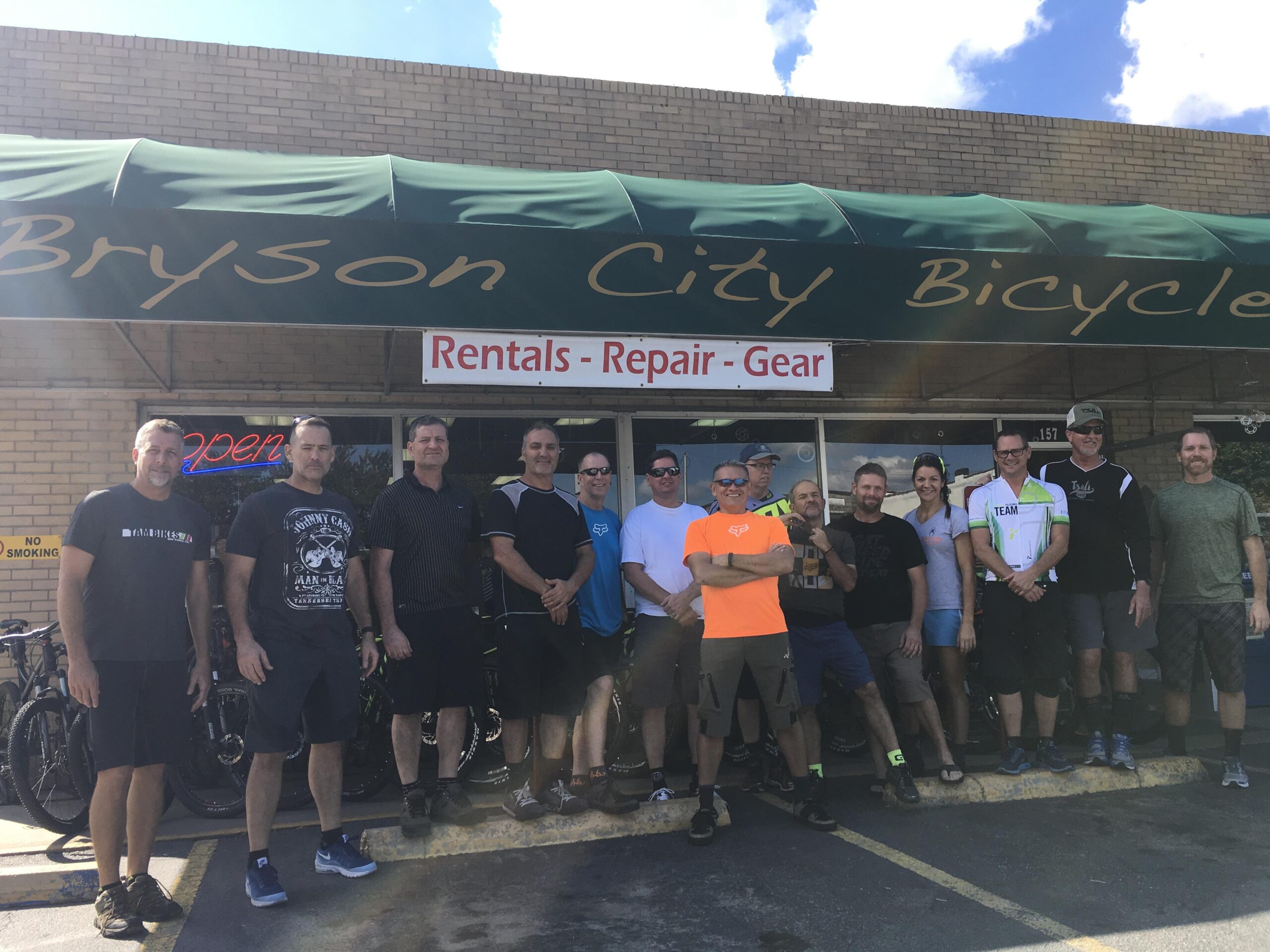 Group photo of a diverse group of individuals standing in front of Bryson City Bicycle, showcasing their shared enthusiasm for cycling. The storefront features a green awning with the shop