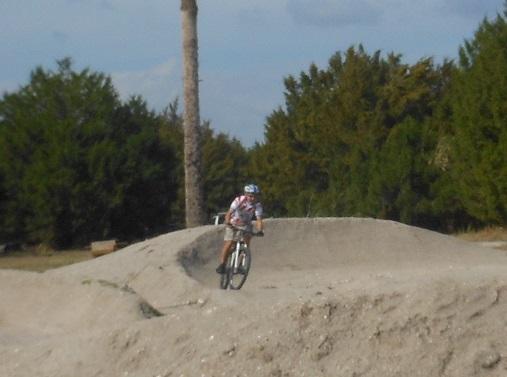 A child in a helmet riding a bicycle on a dirt track with mounds and jumps, surrounded by trees under a cloudy sky. Caloosahatchee Regional Park mountain bike trail.