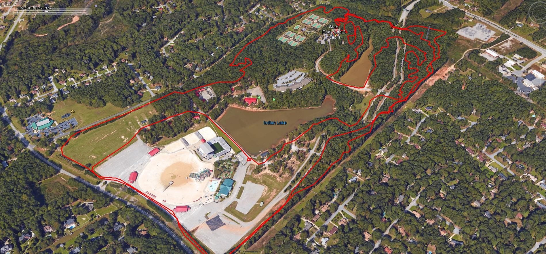 Aerial view of a recreational area featuring a lake labeled "Indian Lake," surrounded by wooded regions and residential neighborhoods. The image includes marked walking or biking trails outlined in red, with areas for activities such as a playground and water park indicated near a central structure. Nearby roads and housing can also be seen in the vicinity. The Trails At The Beach mountain bike trail.
