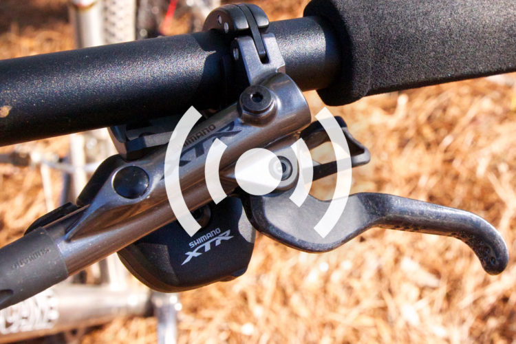 Close-up view of a Shimano XTR bicycle brake lever mounted on a bike handlebar, with a textured grip and a portion of the bike frame visible in the background. The image features a natural outdoor setting with pine needles on the ground.