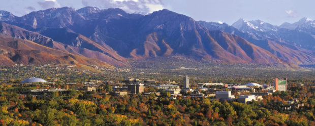 A panoramic view of a university campus surrounded by vibrant autumn foliage, with majestic mountains in the background. The scene captures a mix of greenery and colorful trees, along with various buildings of the campus, highlighting both natural beauty and academic environment. Snow-capped peaks are visible on the horizon under a partly cloudy sky.