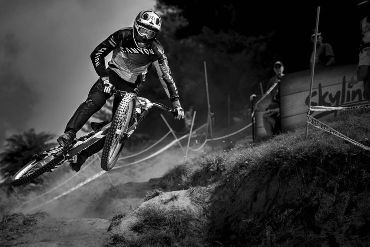 A mountain biker performing a jump on a dirt track, surrounded by dust and spectators in the background. The image is in black and white, highlighting the dynamic movement and intensity of the sport.