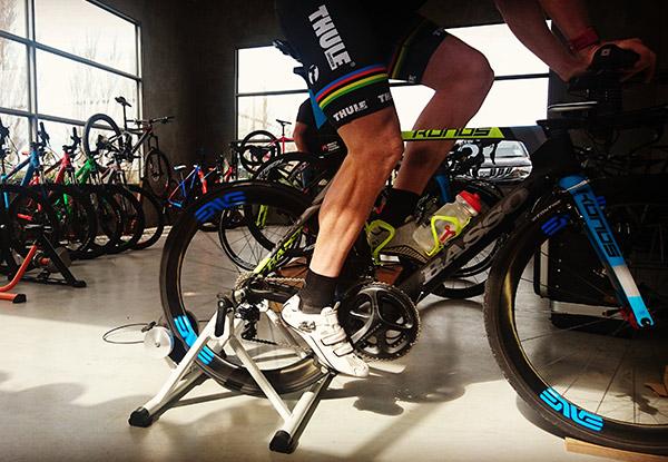 A cyclist wearing shorts and cycling shoes is pedaling on a stationary bike trainer inside a bike shop. The background features various bicycles and bright natural light coming through large windows.