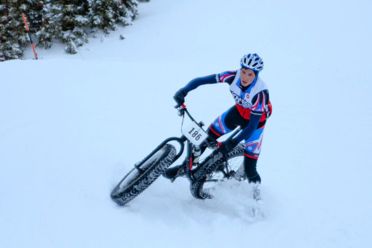 A cyclist dressed in a colorful cycling outfit navigates through deep snow on a mountain bike, with a race number visible on the front. The surrounding landscape is covered in a layer of snow, creating a winter scene.