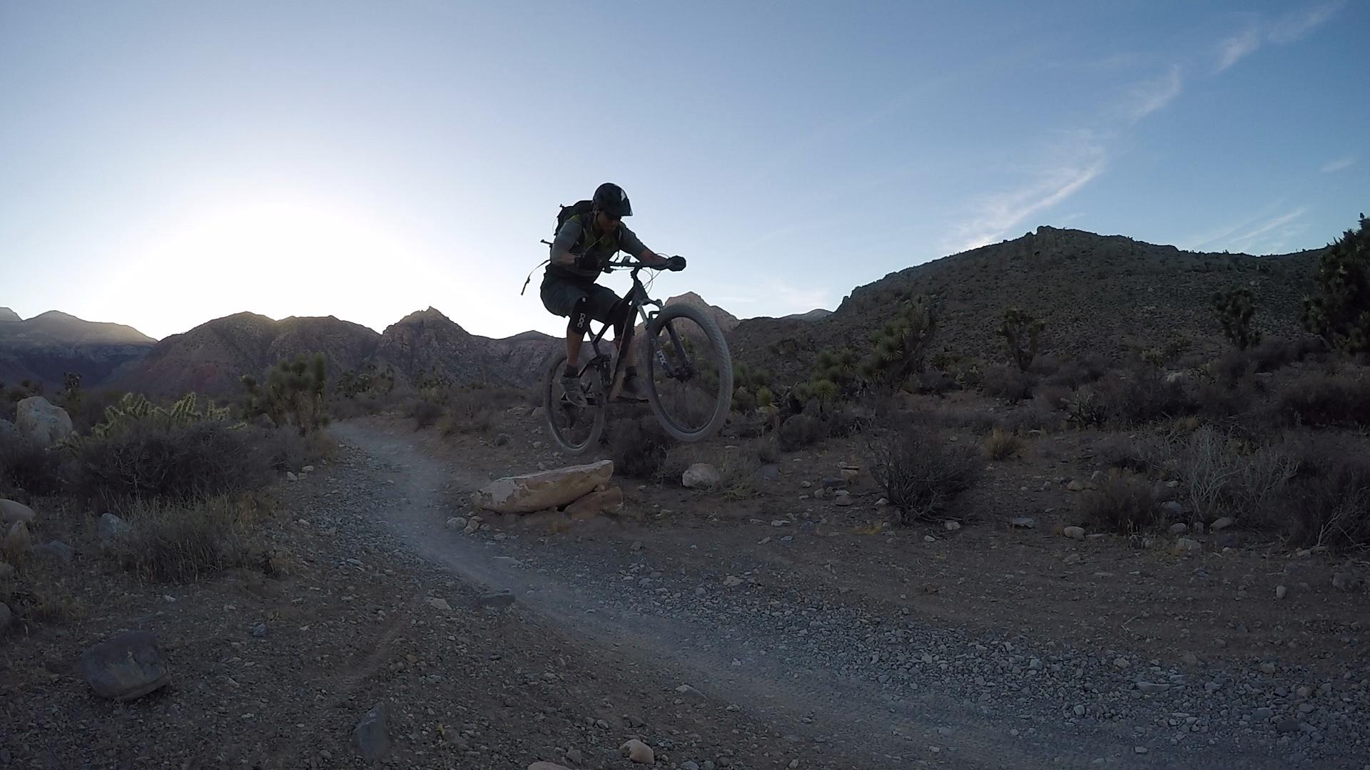 Trek Fuel EX 8: A mountain biker performing a jump over a rock on a dirt path in a desert landscape, with mountains in the background and a clear sky at sunset.