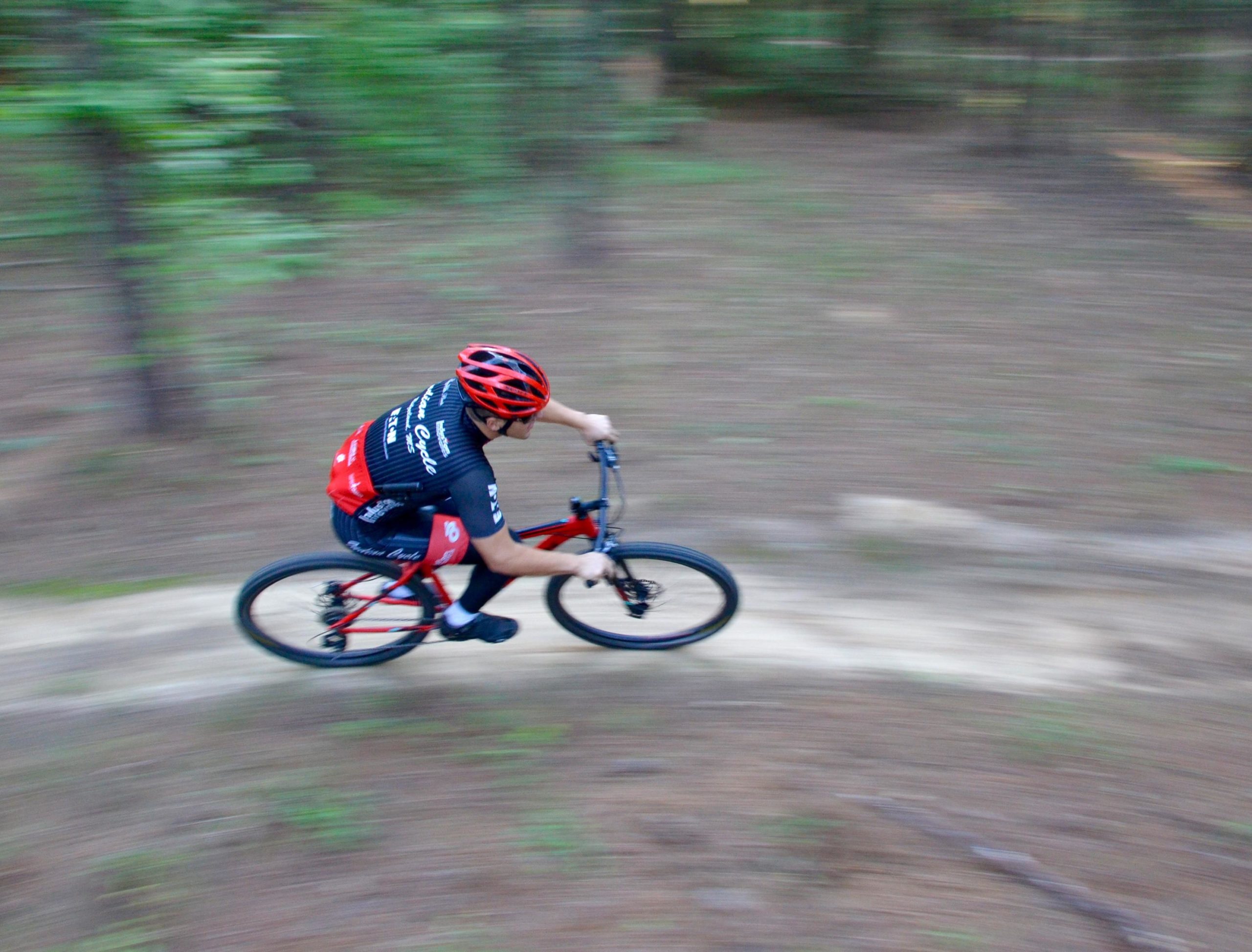 A cyclist in a red and black jersey rides swiftly on a dirt trail through a wooded area, with blurred motion suggesting speed and movement. Mt. Zion Bike Trails mountain bike trail.