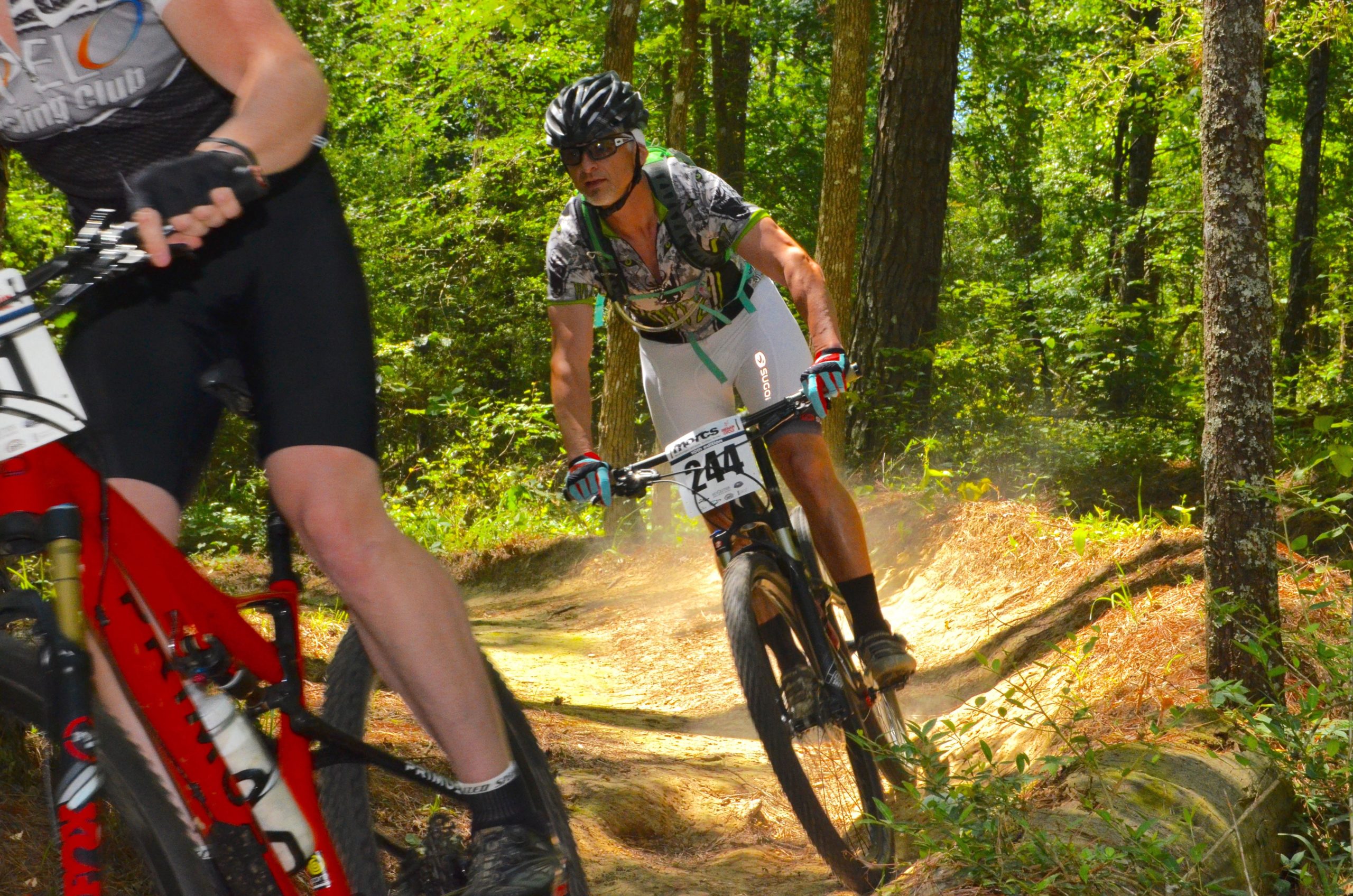 A mountain biker leans into a corner while navigating a dirt trail surrounded by trees, with dust kicked up from the tires. Another cyclist is partially visible in the foreground. Both riders are wearing cycling gear and helmets, indicating an active mountain biking competition or event. Mt. Zion Bike Trails mountain bike trail.