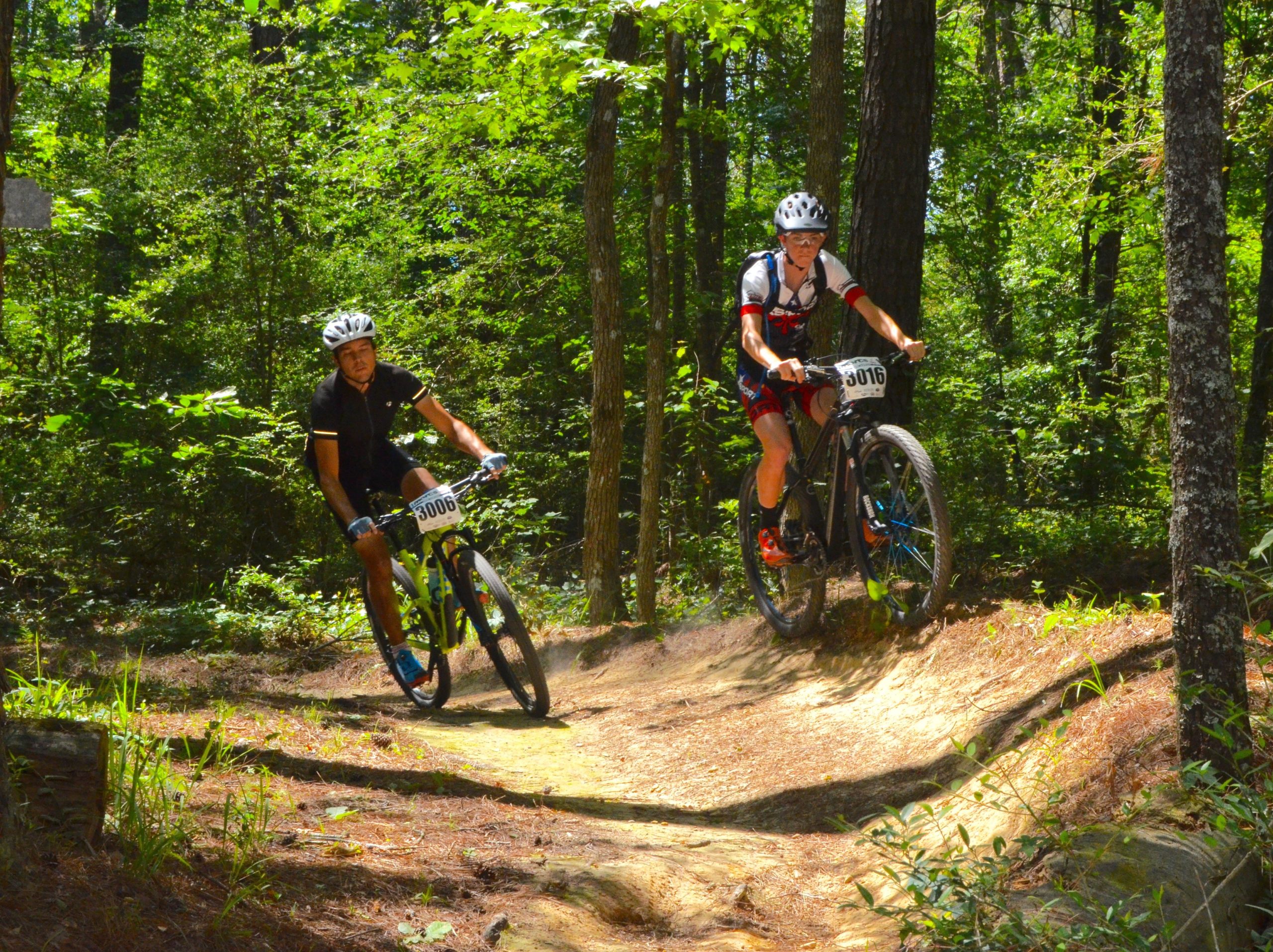 Two mountain bikers navigating a dirt path through a forested area. The rider on the left is dressed in a black jersey with the number 3006, while the rider on the right wears a white jersey with red and blue accents, displaying the number 3016. Sunlight filters through the trees, highlighting the greenery surrounding the trail. Mt. Zion Bike Trails mountain bike trail.