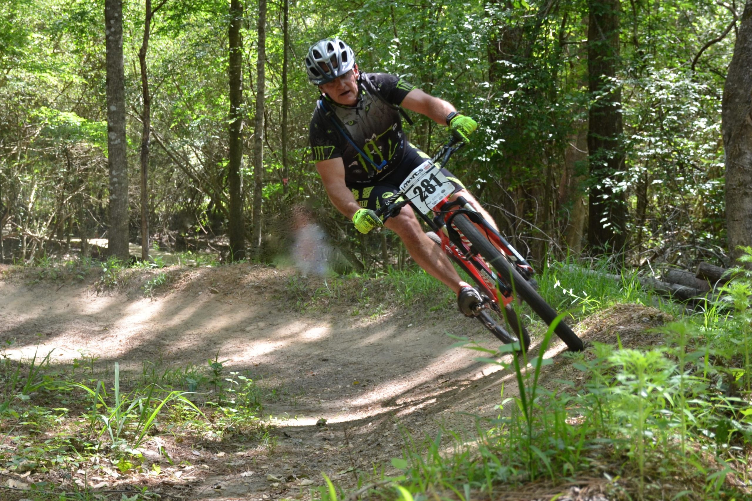 A cyclist navigating a dirt trail in a lush forest, leaning into a turn while riding a mountain bike. The rider is wearing a helmet, gloves, and cycling gear with a race number displayed. Sunlight filters through the trees, illuminating the surrounding greenery and creating a dynamic, active scene. Mt. Zion Bike Trails mountain bike trail.