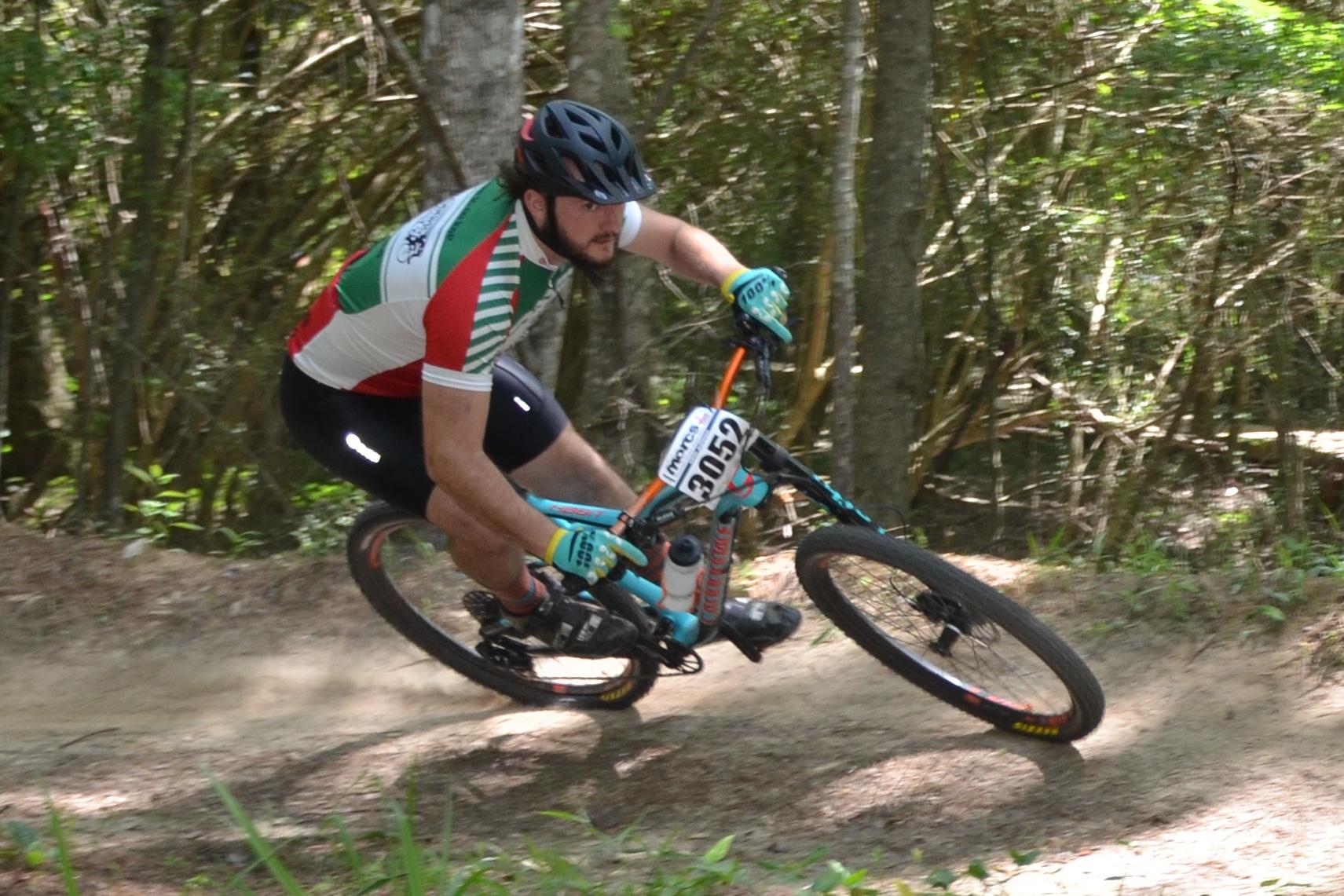 A cyclist in a colorful jersey leans into a turn on a dirt trail, surrounded by trees. Dust is kicked up from the bike as it navigates the rough terrain. The cyclist is wearing a helmet and gloves and is focused on maintaining balance and speed. Mt. Zion Bike Trails mountain bike trail.