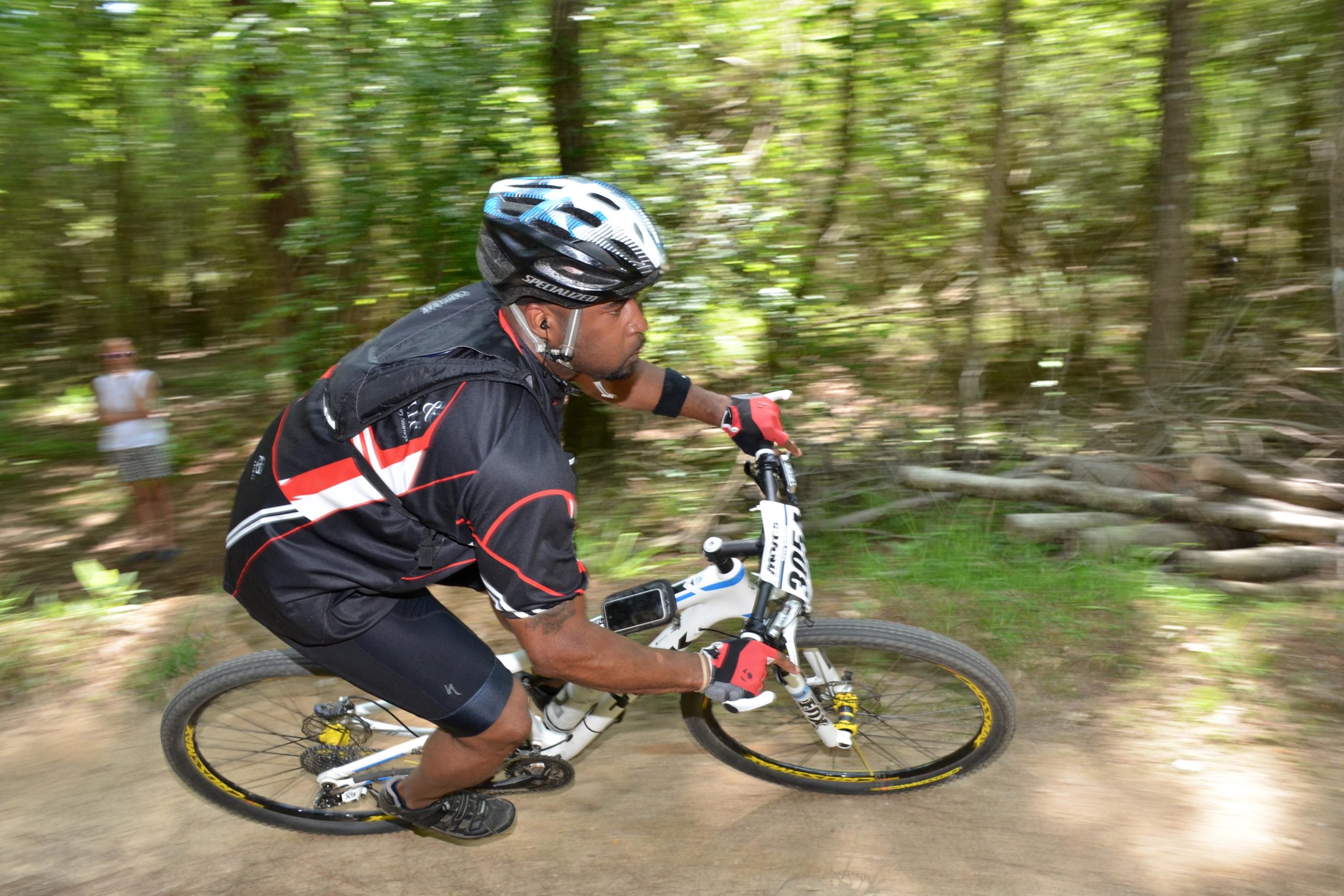 A mountain biker riding on a dirt trail through a wooded area, captured in motion with a blurred background. The biker is wearing a black jersey with red and white accents, a helmet, and gloves, showcasing an intense focus as they navigate the terrain. In the background, a person can be seen observing from a distance. Mt. Zion Bike Trails mountain bike trail.