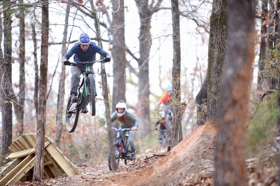 A mountain biker jumps off a wooden ramp in a wooded trail while two other riders, one in the background with a helmet and the other with a beard, are approaching the ramp. The scene captures the excitement of mountain biking through trees and natural terrain. Chewacla State Park mountain bike trail.
