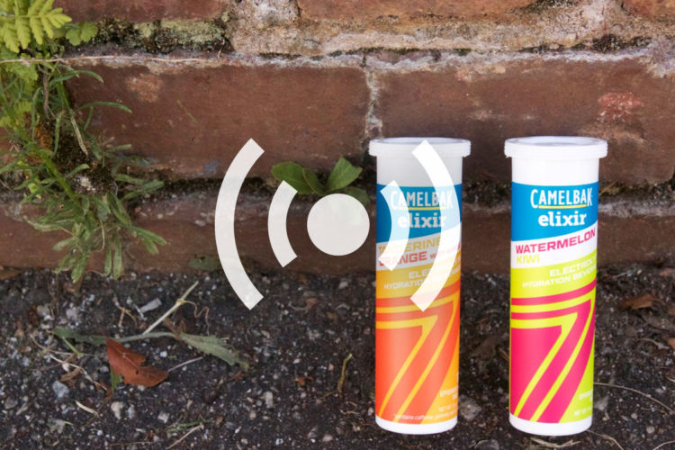 Two tubes of CamelBak Elixir electrolyte hydration tablets are placed next to a brick wall. One tube is labeled "Tangerine Orange," and the other is labeled "Watermelon Kiwi." Green leaves and small plants are visible around the tubes, showcasing a natural setting.