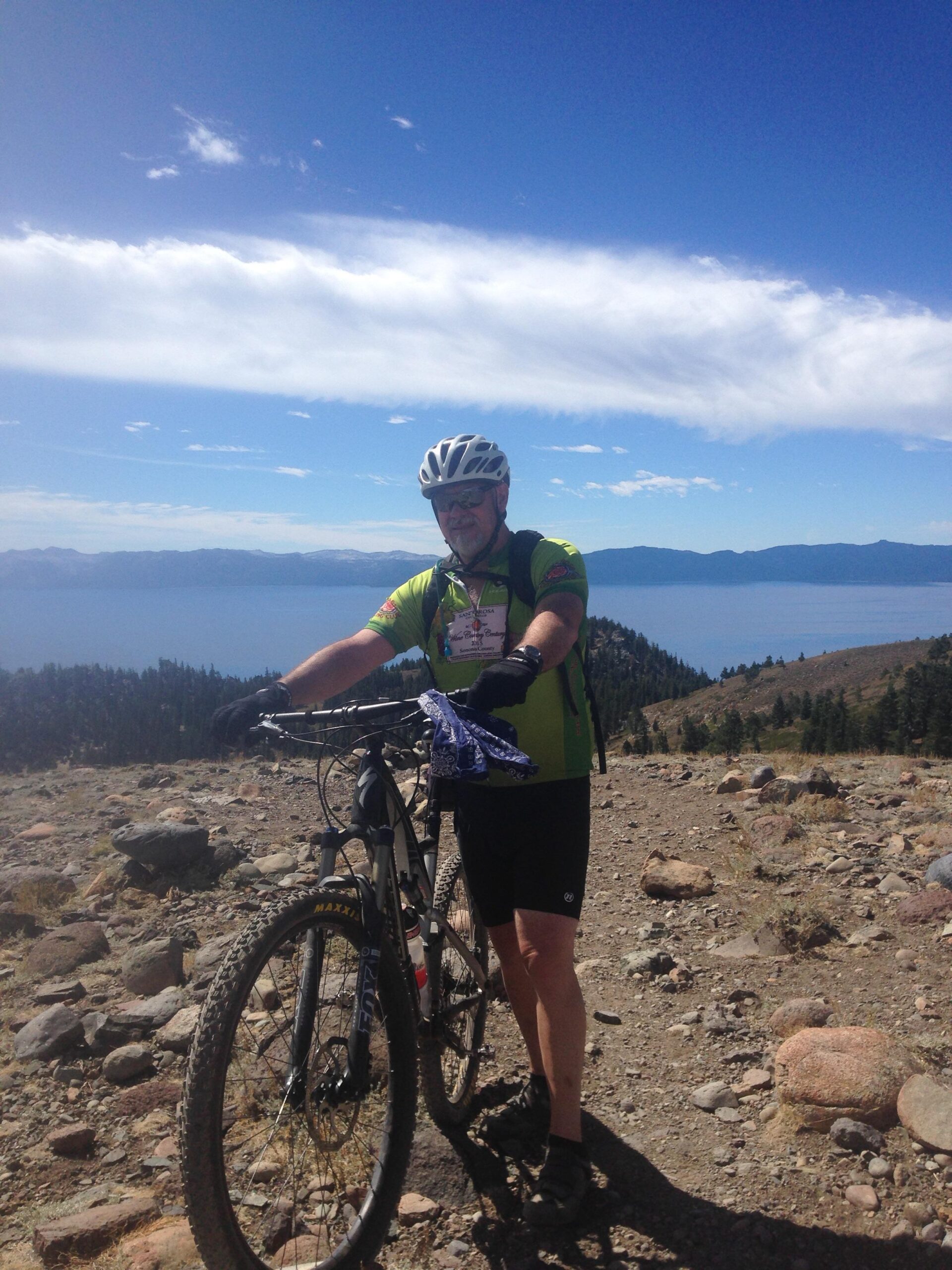 Specialized Camber Comp 29: A mountain biker in a green shirt and cycling shorts stands beside his bike on a rugged trail with rocky terrain. In the background, a serene blue lake stretches out under a clear sky with scattered clouds and distant mountain ranges. The scene captures the beauty of outdoor adventure and the thrill of mountain biking.