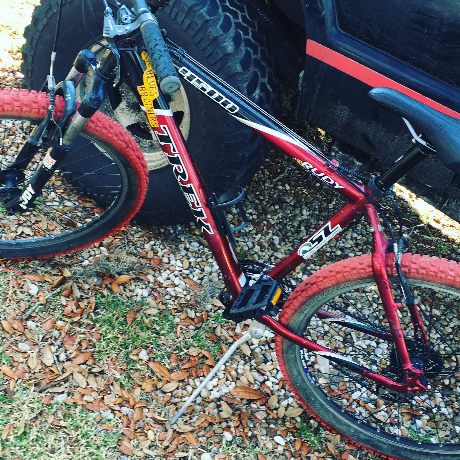Trek 4500: A red Trek mountain bike parked on gravel and grass, with a large tire of a vehicle visible in the background. The bike features thick, knobby tires and a sturdy frame, suggesting it’s designed for off-road use. Dry leaves are scattered on the ground around it.