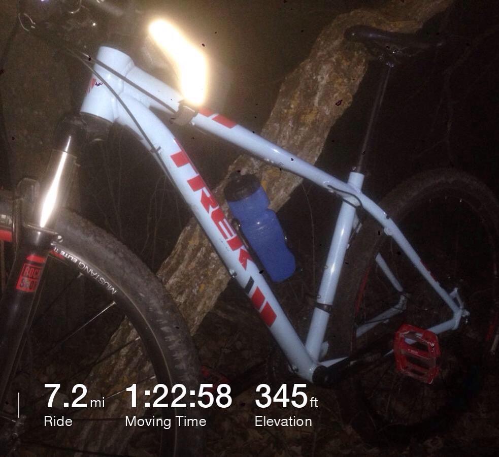 Trek X-Caliber: A close-up image of a Trek mountain bike resting against a tree in a dark setting. The bike features a light reflecting off its frame and has a blue water bottle attached to it. A digital display in the foreground shows the ride statistics: 7.2 miles traveled, a moving time of 1:22:58, and an elevation gain of 345 feet.