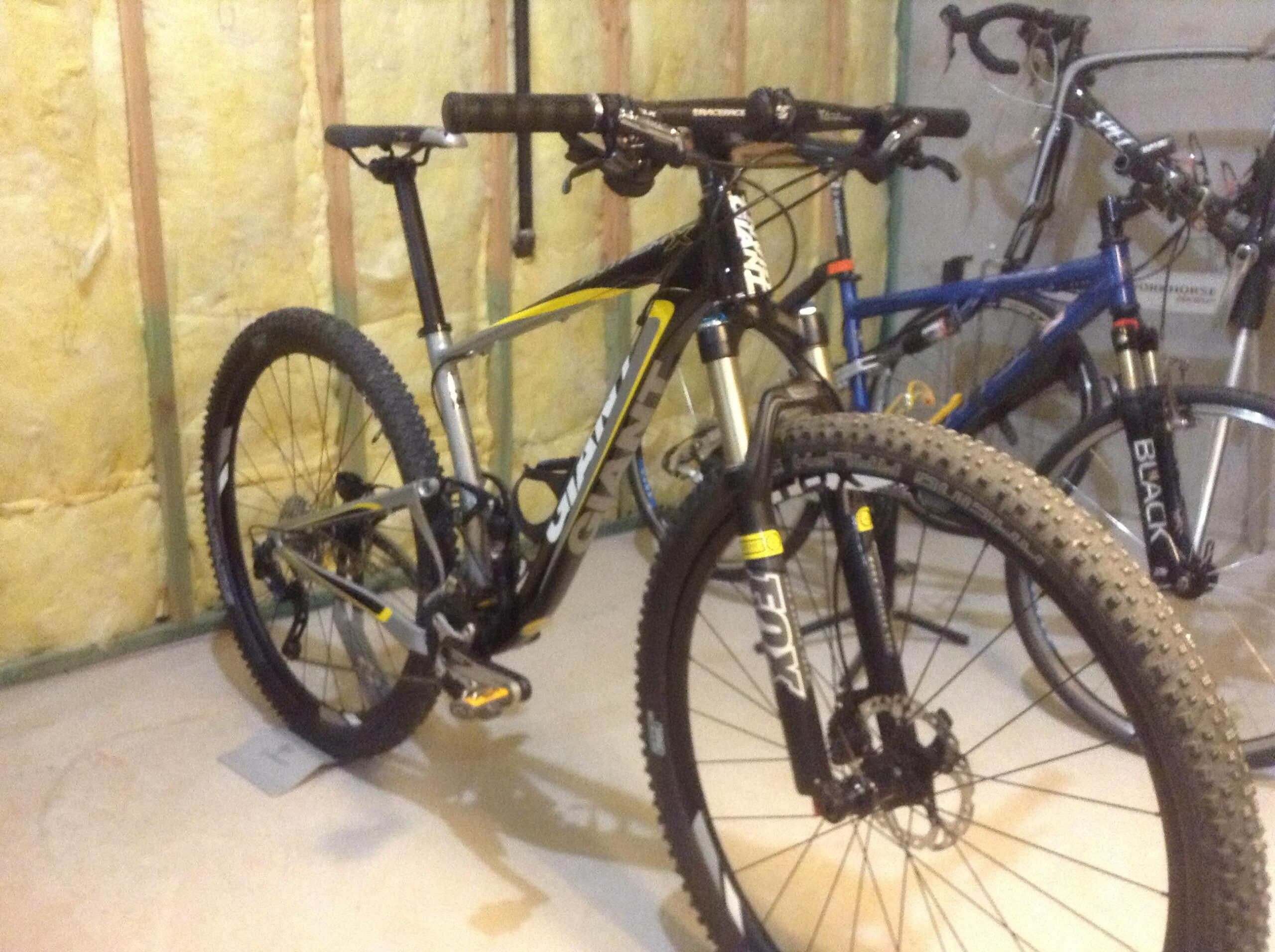 Giant Anthem: A mountain bike with a sleek black and yellow frame is positioned in a garage. The bike features thick, textured tires suitable for off-road terrain and front suspension forks. In the background, another bike is partially visible, along with bare walls insulated with yellow material, indicating a garage or storage space.