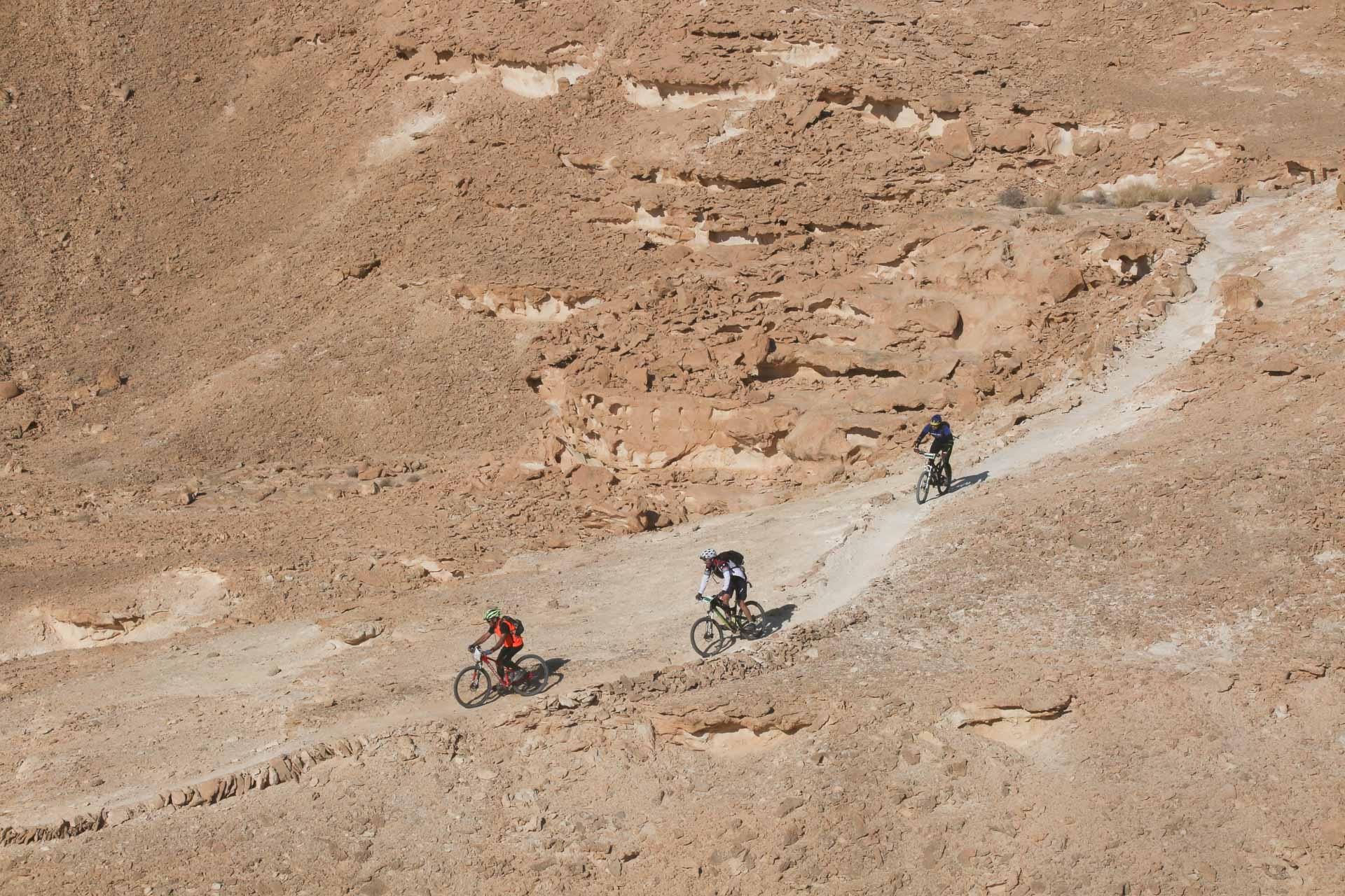 Santa Cruz Heckler: Three mountain bikers navigate a rugged dirt trail in a desert landscape, surrounded by rocky hills and dry terrain under a clear sky.