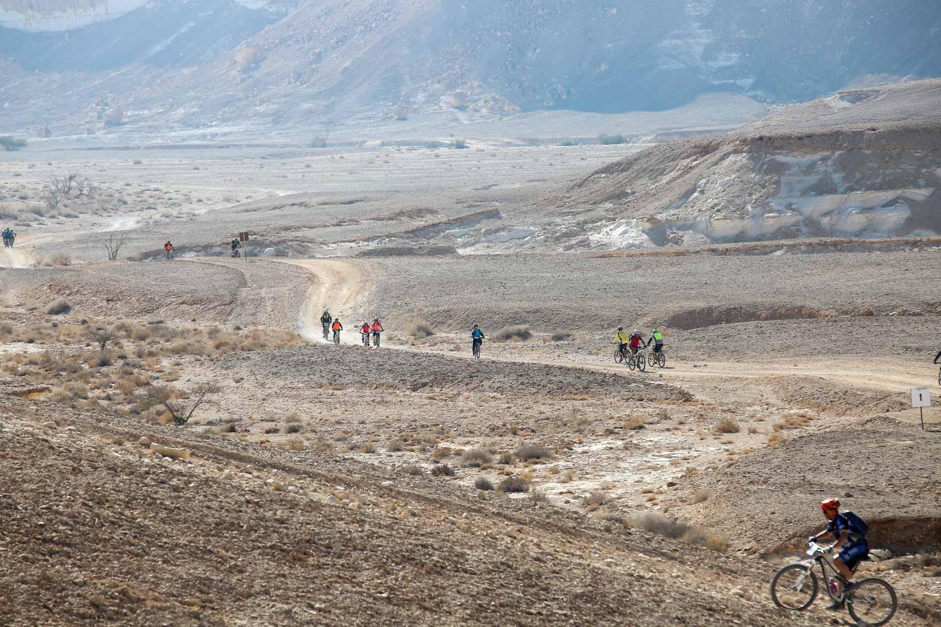 Santa Cruz Heckler: A group of cyclists riding on a dirt path through a sparse, rugged landscape with rocky hills in the background. The scene features several individuals, some wearing bright clothing, as they traverse the hilly terrain under a clear sky.
