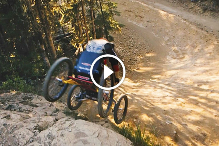 A person in a specialized off-road wheelchair is jumping over a rocky terrain on a dirt path surrounded by trees. The action captures the thrill of extreme sports, showcasing the wheelchair's capability on rugged trails.