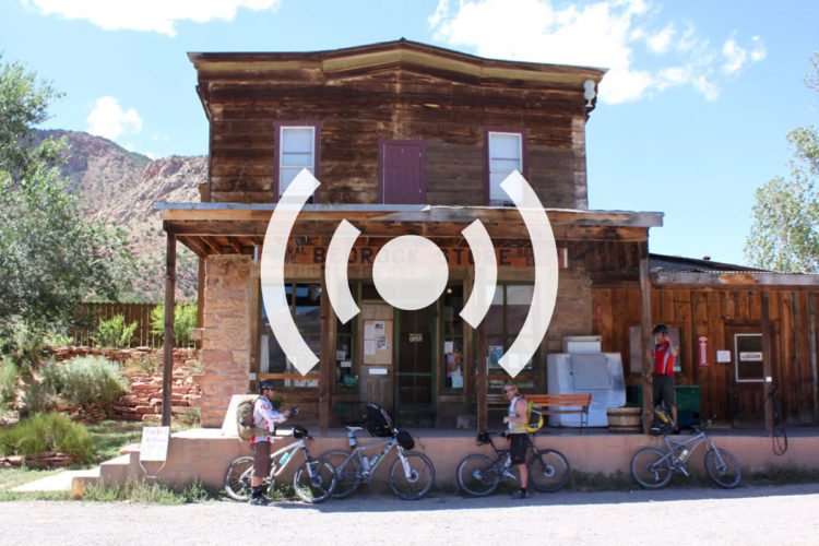 A rustic two-story wooden building labeled "Bedrock Store" is depicted with cyclists standing next to their bikes outside the entrance. The surrounding area features greenery and red rock formations under a blue sky with scattered clouds.