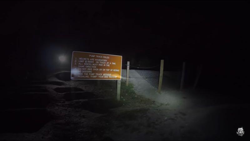 A dimly lit pathway at night, featuring a sign with rules for a pump track. The sign is illuminated by a flashlight and displays guidelines such as "Helmets are mandatory" and "Only one rider at a time," surrounded by a fenced area. The background is dark, with outlines of terrain visible. Jonathan Dickinson State Park mountain bike trail.