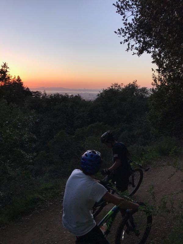 Two mountain bikers pause on a dirt trail, overlooking a scenic view as the sun sets in the background, painting the sky with shades of orange and blue. The surrounding landscape is lush with trees, providing a natural backdrop to their outdoor adventure. Joaquin Miller mountain bike trail.