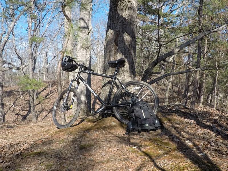 A black mountain bike with a helmet attached is resting against a tree in a wooded area. Nearby, there is a black backpack on the ground, surrounded by dry leaves and moss. The scene is set in a sunny outdoor environment with trees and blue sky visible in the background. Chicopee Woods mountain bike trail.