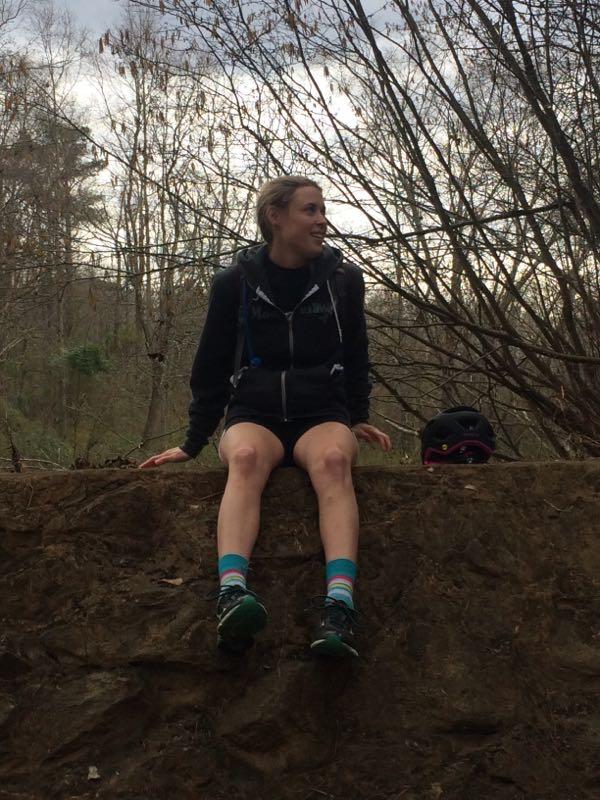 A person sitting on a rocky outcrop in a wooded area, wearing a black jacket and shorts, with colorful socks and hiking shoes. There is a helmet resting beside them, and the background features bare trees and a cloudy sky. Harris Shoals mountain bike trail.