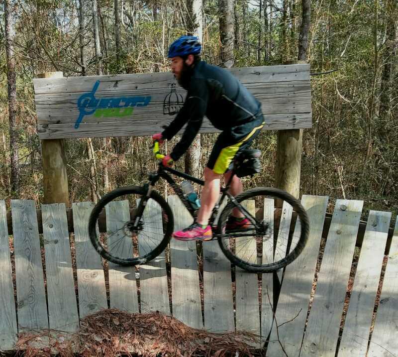 A mountain biker in a black jacket and yellow shorts jumps over a wooden fence near a trail entrance sign in a wooded area. The sign features the words "Cycling Trail" and is surrounded by trees and natural foliage. Golden Eagle Trail Complex / Tuffburg / Longleaf Trace mountain bike trail.