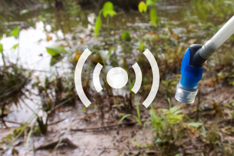 A close-up of a water sampling device positioned near a muddy bank, with a blurred background of water and lush green vegetation. The device features a blue connector and a transparent tube, suggesting it is designed for collecting or monitoring water quality. A graphic symbol indicating connectivity or transmission overlays the image.