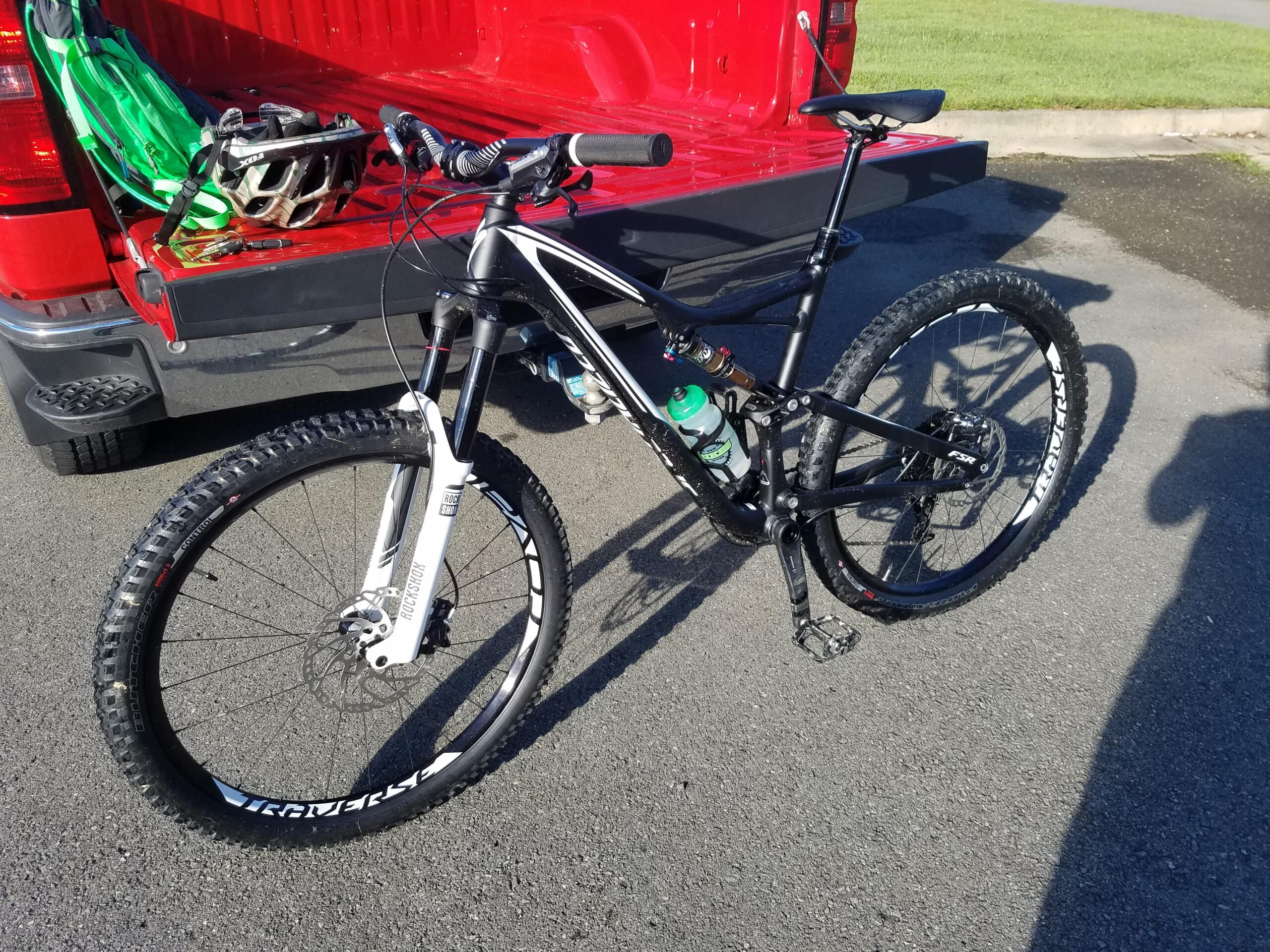 Specialized Stumpjumper FSR Expert: A black and white mountain bike parked next to the tailgate of a red truck. The bike features thick tires and a water bottle attached to the frame. In the background, a green backpack and a helmet can be seen on the truck bed. The setting appears to be an outdoor area with grass in the distance.