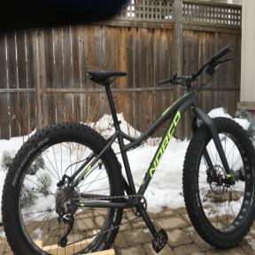 Norco Bigfoot: A black and green fat bike featuring wide tires, parked on a snow-covered surface with a wooden fence in the background.