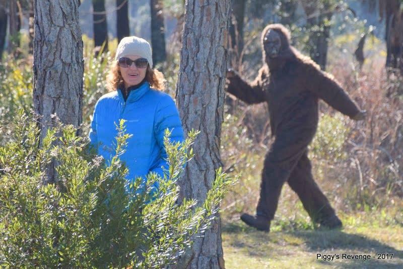 A woman wearing a blue jacket and sunglasses stands next to a tree in a wooded area, looking surprised. In the background, a person in a Sasquatch costume walks through the forest. The scene captures a playful interaction between the woman and the costumed figure. BoldlyGo mountain bike trail.