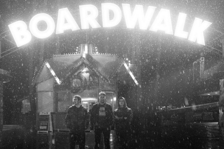 A black and white image of three people standing under a large illuminated sign that reads "BOARDWALK." The scene is set in a rainy environment with falling raindrops visible, and the background features a building with festive lights, indicative of an amusement or entertainment area.