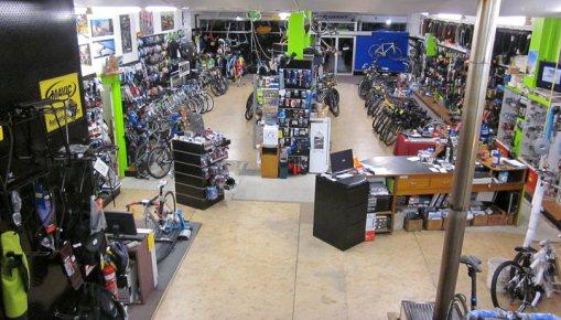 A spacious bike shop filled with various bicycles, bike accessories, and equipment. The interior features a central sales counter and numerous display racks showcasing different bike models and gear, with bright green accent walls. The layout is open, allowing for easy navigation between the aisles of bicycles.