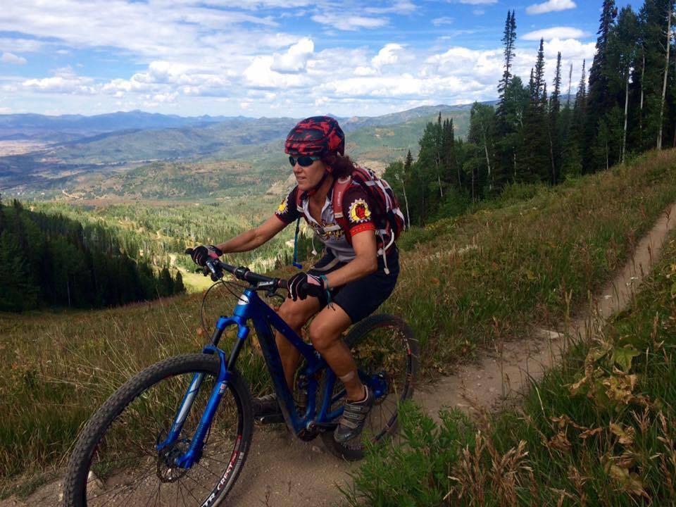 Niner R.I.P. 9 RDO: A person riding a mountain bike on a scenic trail surrounded by green grass and trees, with a panoramic view of mountains in the background under a partly cloudy sky. The cyclist is wearing a helmet and a cycling jersey, and a backpack is attached to their back.