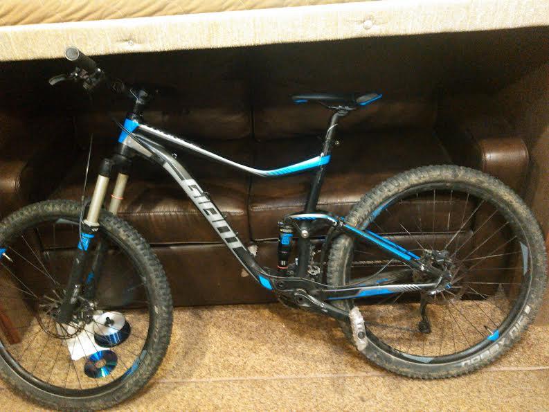 Giant Trance 27.5 3: A mountain bike with a black and blue frame, featuring large tires and a front suspension fork, is leaning against a brown leather couch. The bike's tires show signs of dirt, indicating recent use. A blue water bottle is attached to the bike frame.