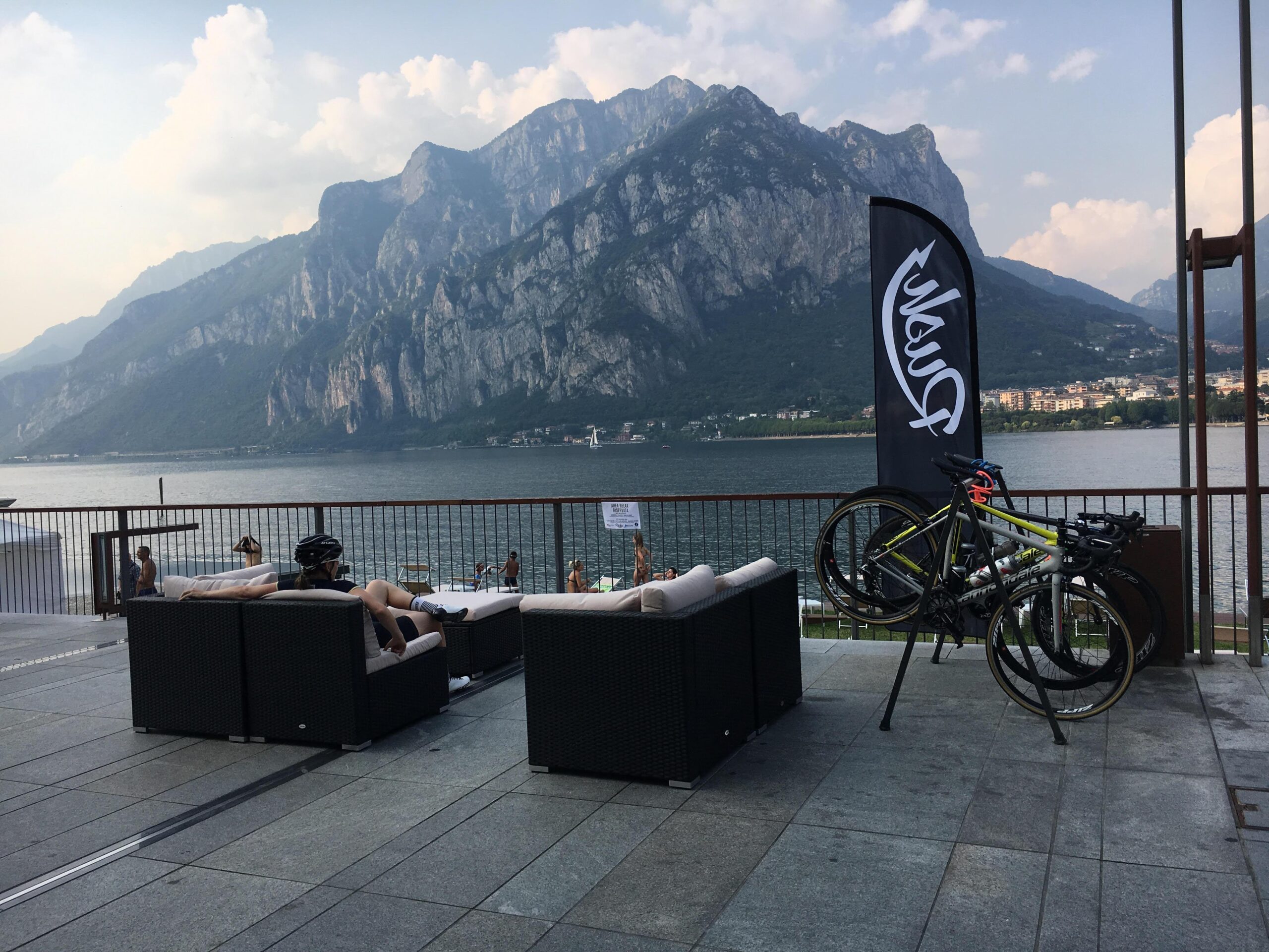 A scenic lakeside view featuring a cyclist relaxing on a lounge chair with a helmet beside them, while another person swims nearby. In the foreground, there are several modern lounge chairs and two bicycles secured on a bike rack. A large flag with the logo "Puma" stands prominently. The backdrop showcases majestic mountains and a calm lake under a partly cloudy sky.