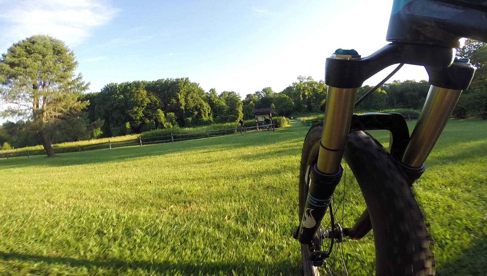 A close-up view of a mountain bike's front fork and tire on a grassy path, with a lush green landscape in the background. A wooden structure can be seen in the distance, surrounded by trees under a clear blue sky. Huber Woods mountain bike trail.