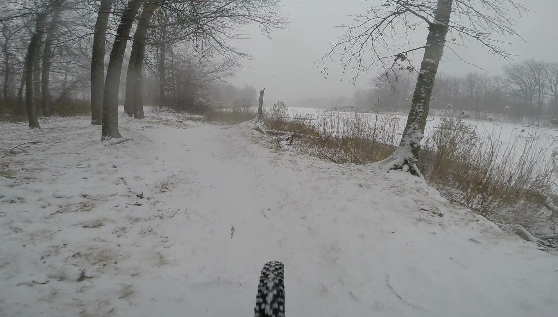 A snowy bike trail winds through a forest in winter, with snow-covered ground and trees partially obscured by swirling snowflakes. In the foreground, a bicycle tire is visible, indicating motion along the path. The scene is gray and misty, capturing the serene beauty of a snow-covered landscape. Wolfes Pond park mountain bike trail.