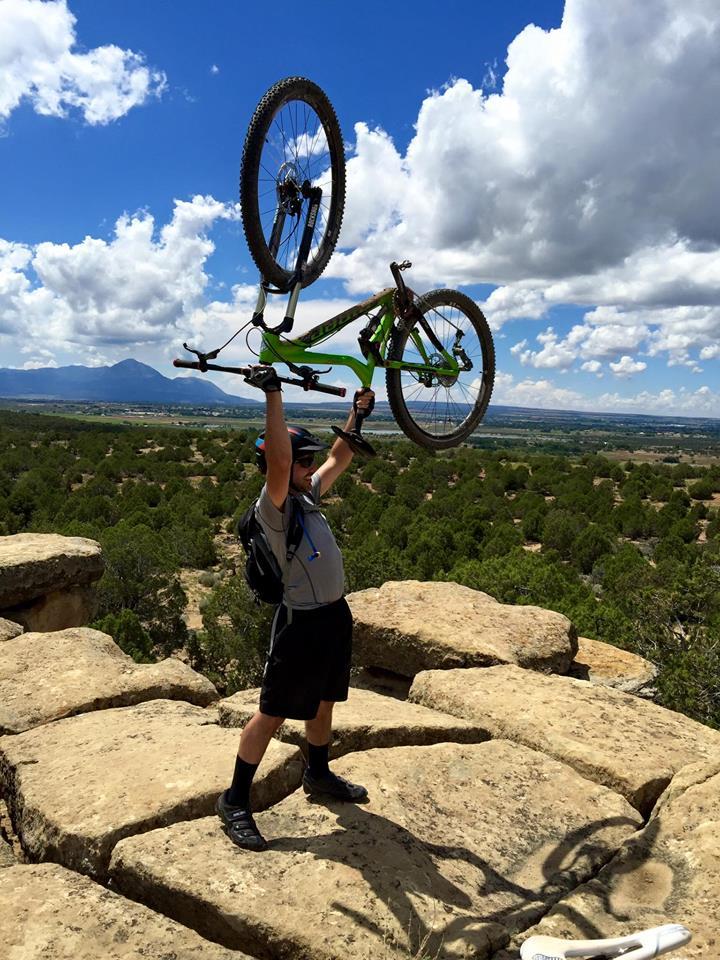 Niner RIP 9: A person stands on rocky terrain, triumphantly holding a mountain bike overhead with both hands. The background features a scenic view of rolling hills and a blue sky dotted with fluffy white clouds. The individual is dressed in athletic gear, including a helmet and a backpack, indicating an outdoor adventure.