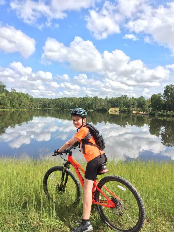 A child wearing a helmet and an orange shirt stands beside a red mountain bike on a grassy bank, looking back at the camera. In the background, a calm lake reflects the blue sky and fluffy white clouds, surrounded by greenery and trees. Colonel Francis Beatty Park mountain bike trail.