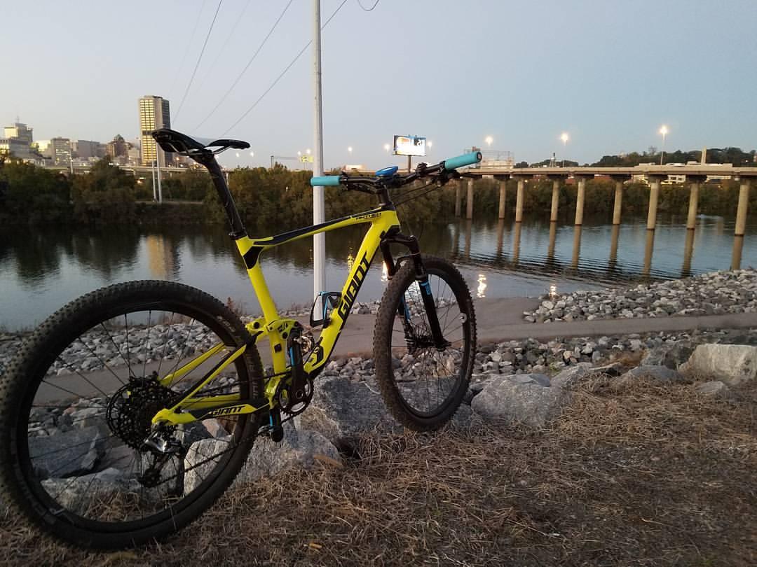 Giant Anthem Advanced 27.5 1: A bright yellow mountain bike is positioned on rocky terrain near a riverbank, with a city skyline in the background. The scene is set during dusk, with reflections on the water and city lights starting to illuminate the skyline and bridge.