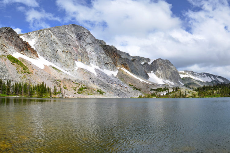 A panoramic view of a serene alpine lake surrounded by rugged mountains. The mountains are partially covered in snow, contrasting with the green vegetation and rocky terrain. Soft clouds float in a blue sky, reflecting on the calm water's surface.