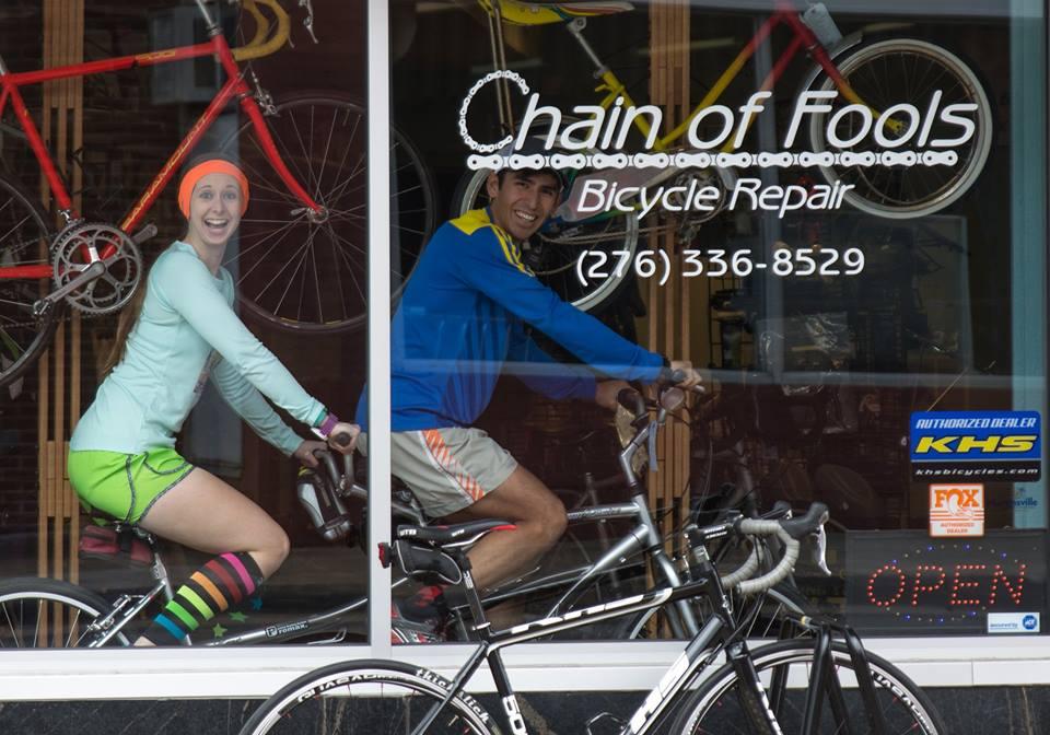 Two smiling individuals are sitting on bicycles inside a bicycle repair shop, "Chain of Fools." The woman, wearing a bright orange headband and colorful striped socks, is positioned on the left, while the man, dressed in a blue and yellow athletic outfit, is on the right. In the background, various bicycles are displayed, and the shop