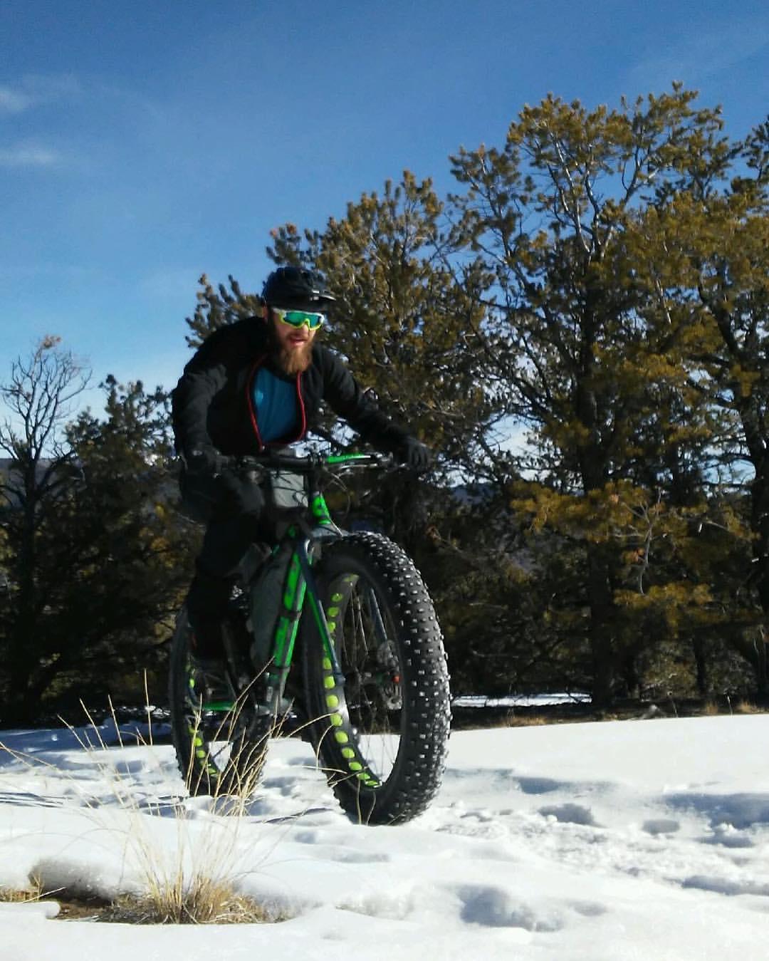 A person riding a fat tire bicycle through a snowy landscape, surrounded by trees under a clear blue sky. The cyclist has a beard, wears a helmet and sunglasses, and is dressed in warm outdoor gear. Little Rainbow mountain bike trail.