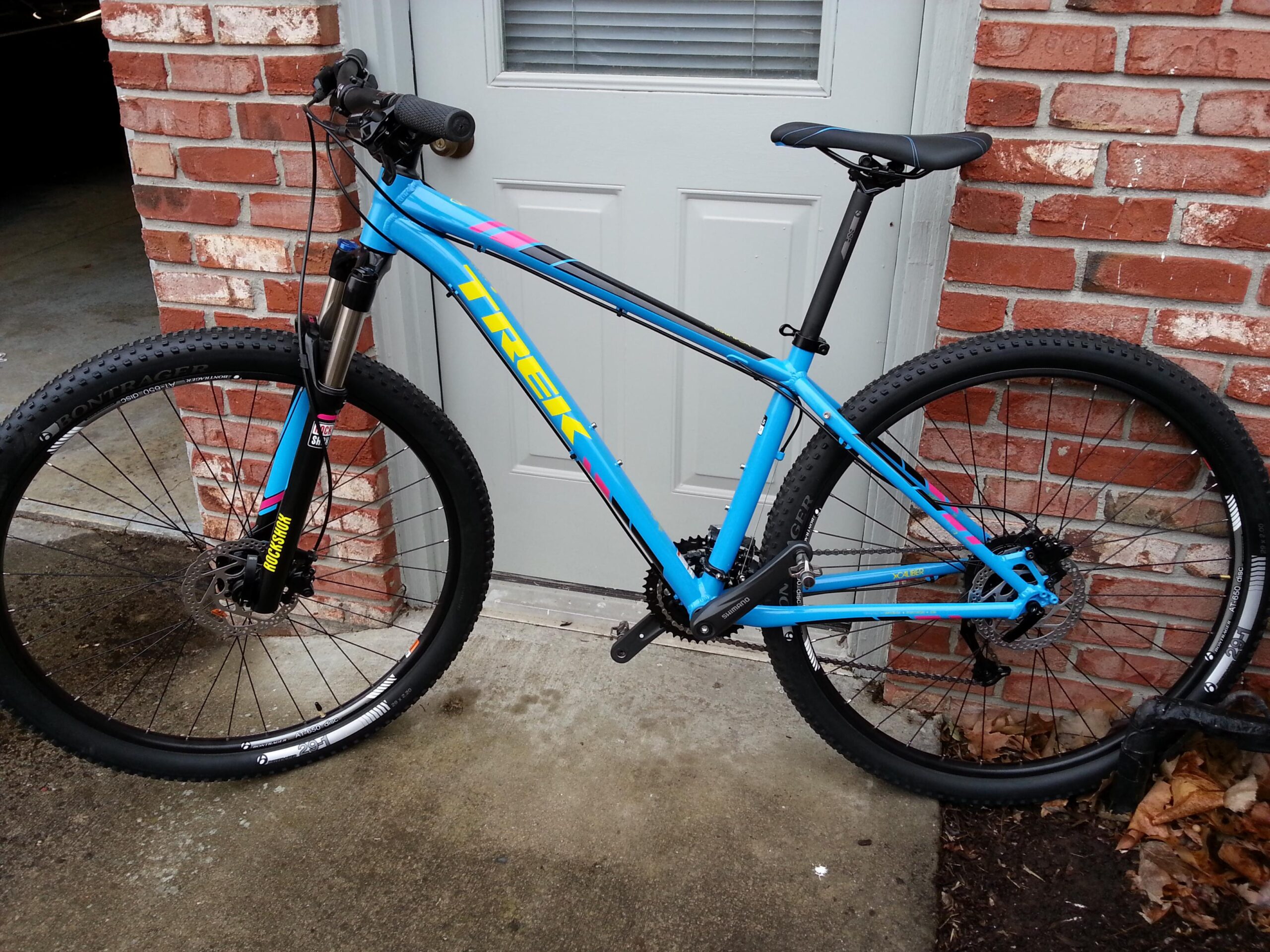 Trek X-Caliber 8: A blue Trek mountain bike with front suspension, positioned next to a brick wall and a door. The bike features thick tires, disc brakes, and a sleek design suitable for off-road riding. Brown leaves are scattered on the ground nearby.