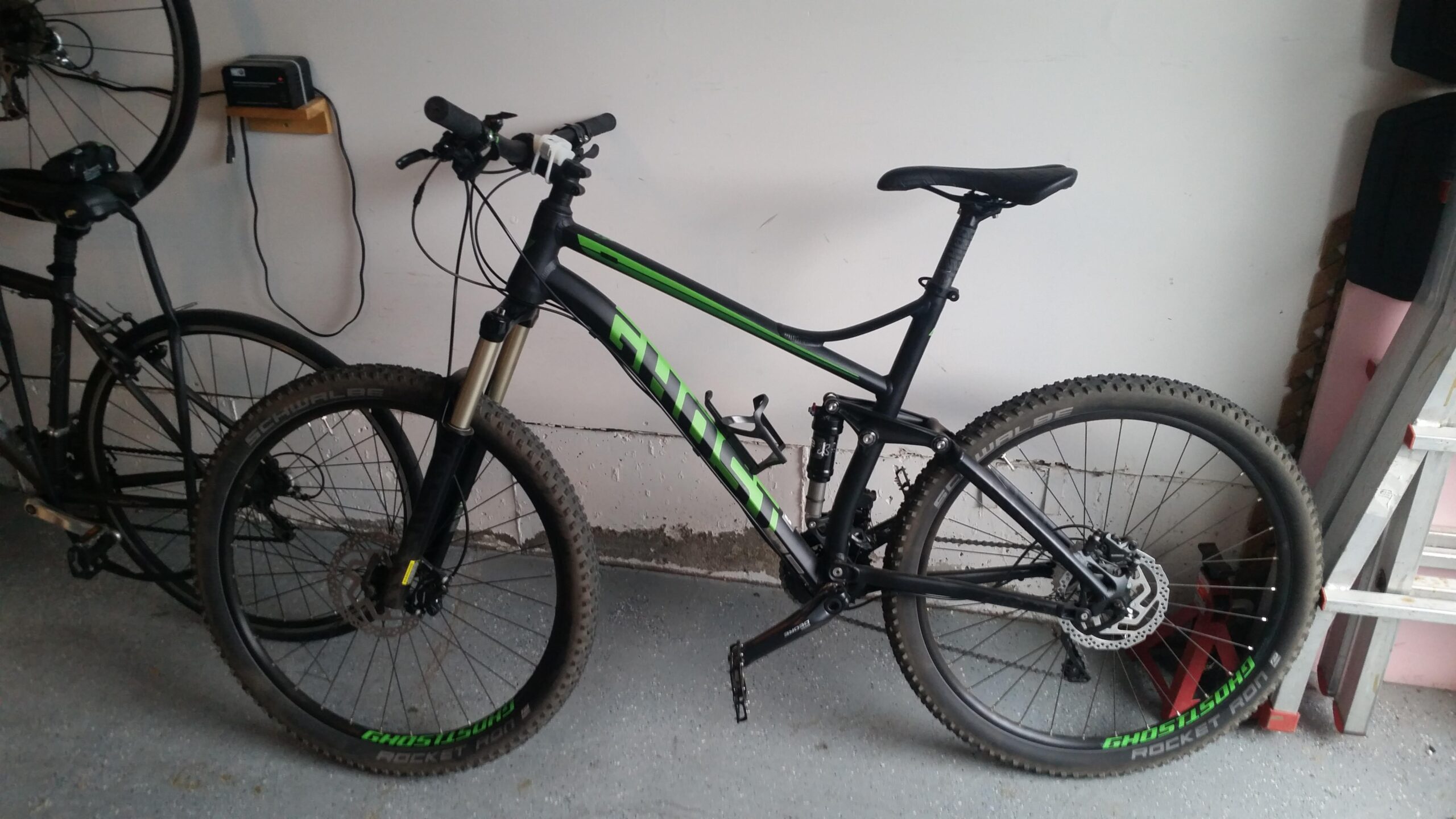 Ghost Kato 3: Alt text: A black and green mountain bike is leaning against a wall in a garage, next to another bike. The bike features thick tires with visible tread, front suspension, and a water bottle holder. The garage has a simple concrete floor and some tools and other items in the background.